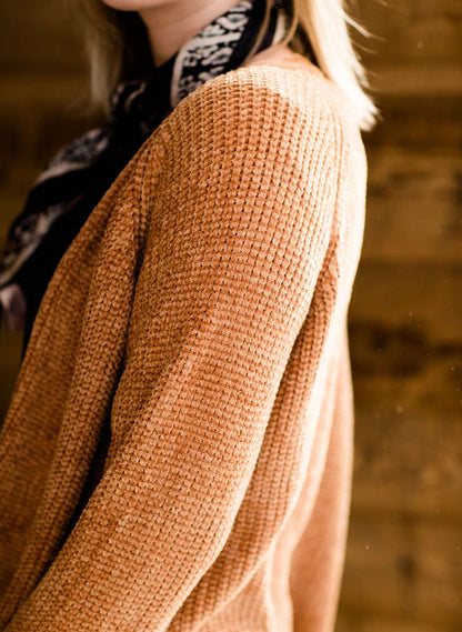 Woman wearing an open front cardigan that is chenille and camel in color. She is also wearing it over a black dress and brown riding boots. She is standing on the stairs at Inherit Clothing Company.