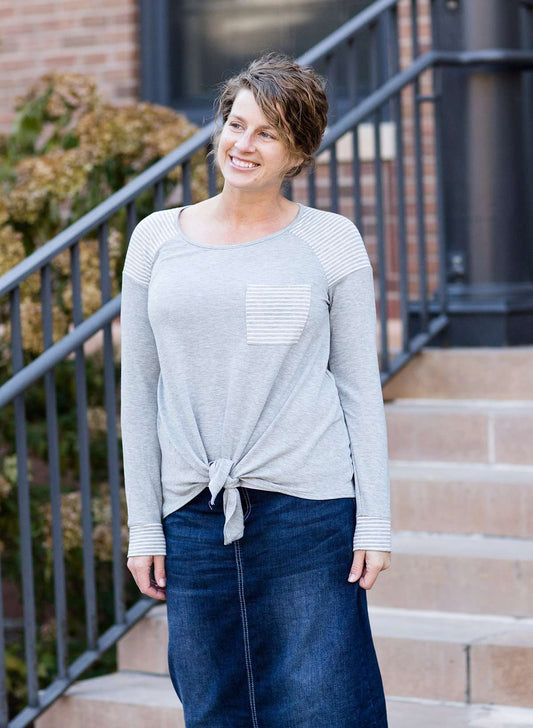 Woman wearing a gray tri blend top that has a striped pocket and a front tie.