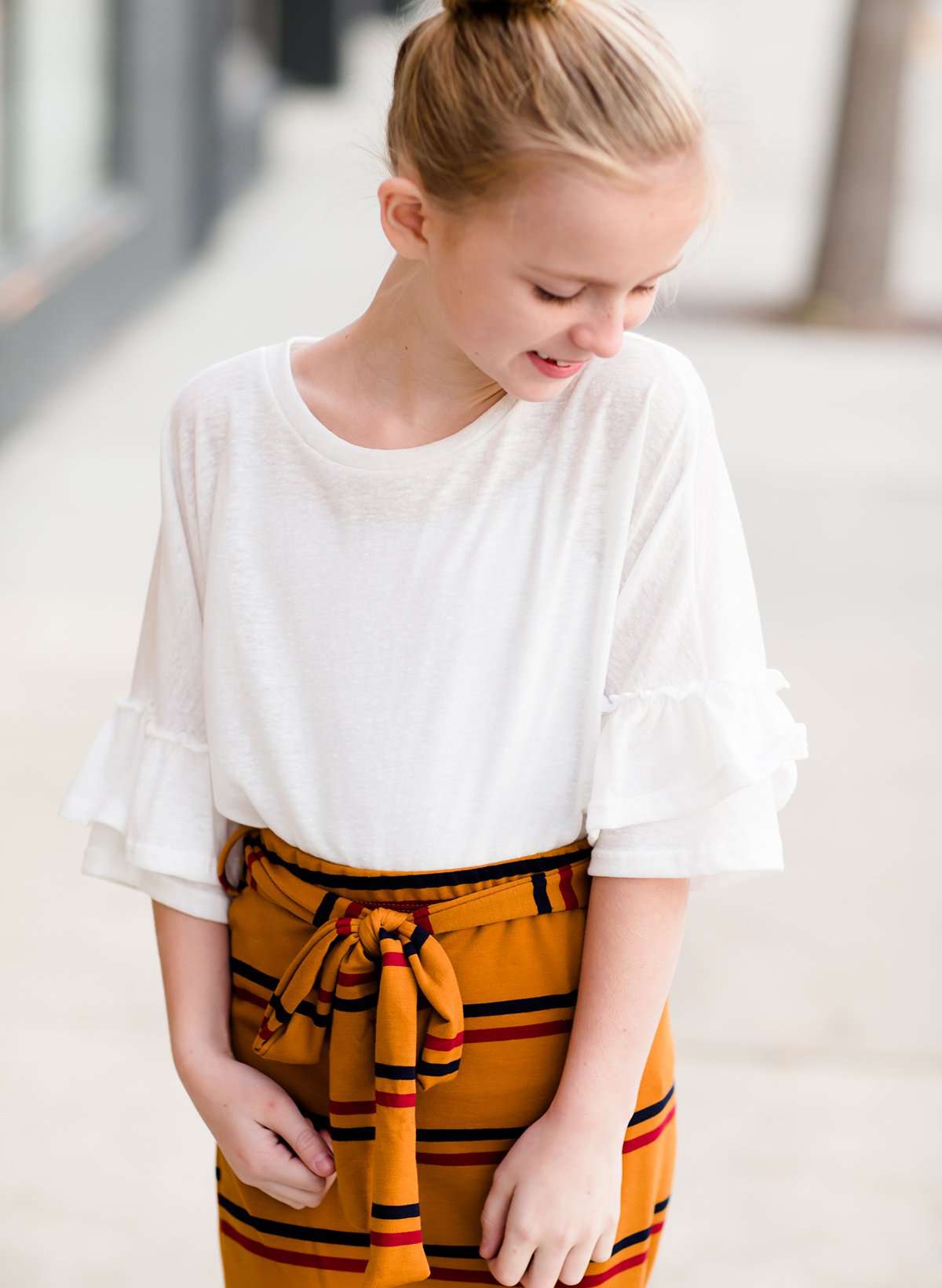 Young girl wearing an ivory, oversized modest top with ruffle sleeve accents. This top is also paired with a mustard, striped below the knee skirt and short boots.