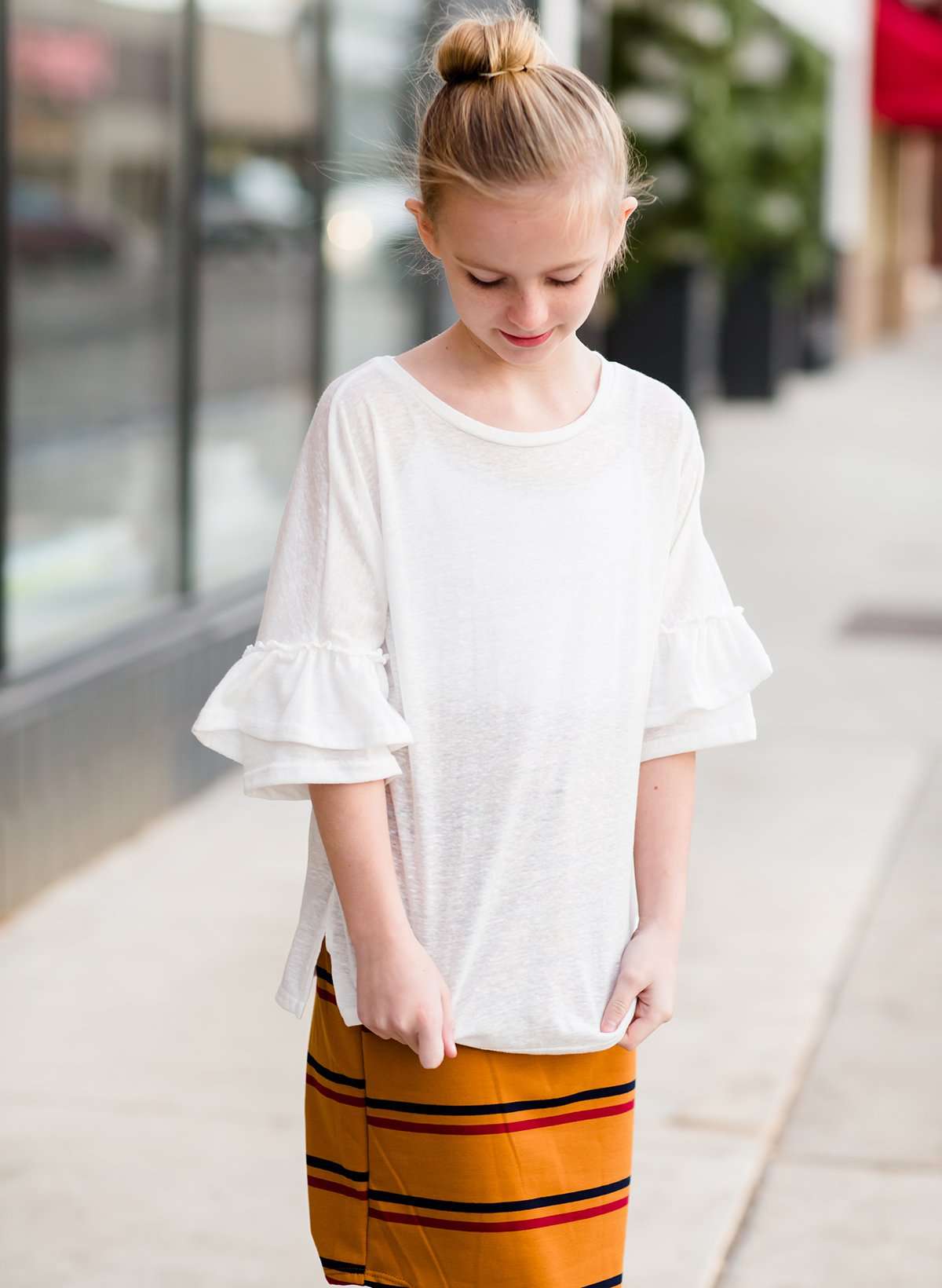 Young girl wearing an ivory, oversized modest top with ruffle sleeve accents. This top is also paired with a mustard, striped below the knee skirt and short boots.