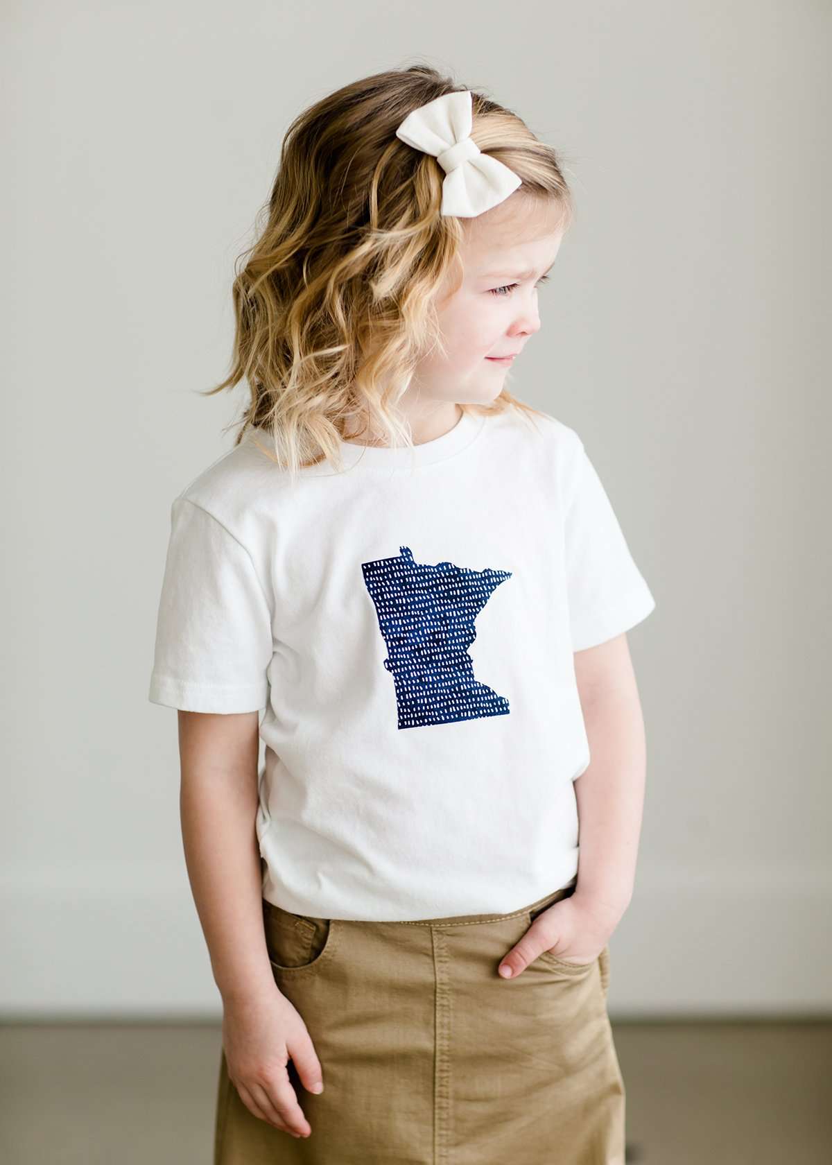 Toddler girl wearing a white minnesota logo on a white tee shirt. She is also wearing a long khaki skirt.