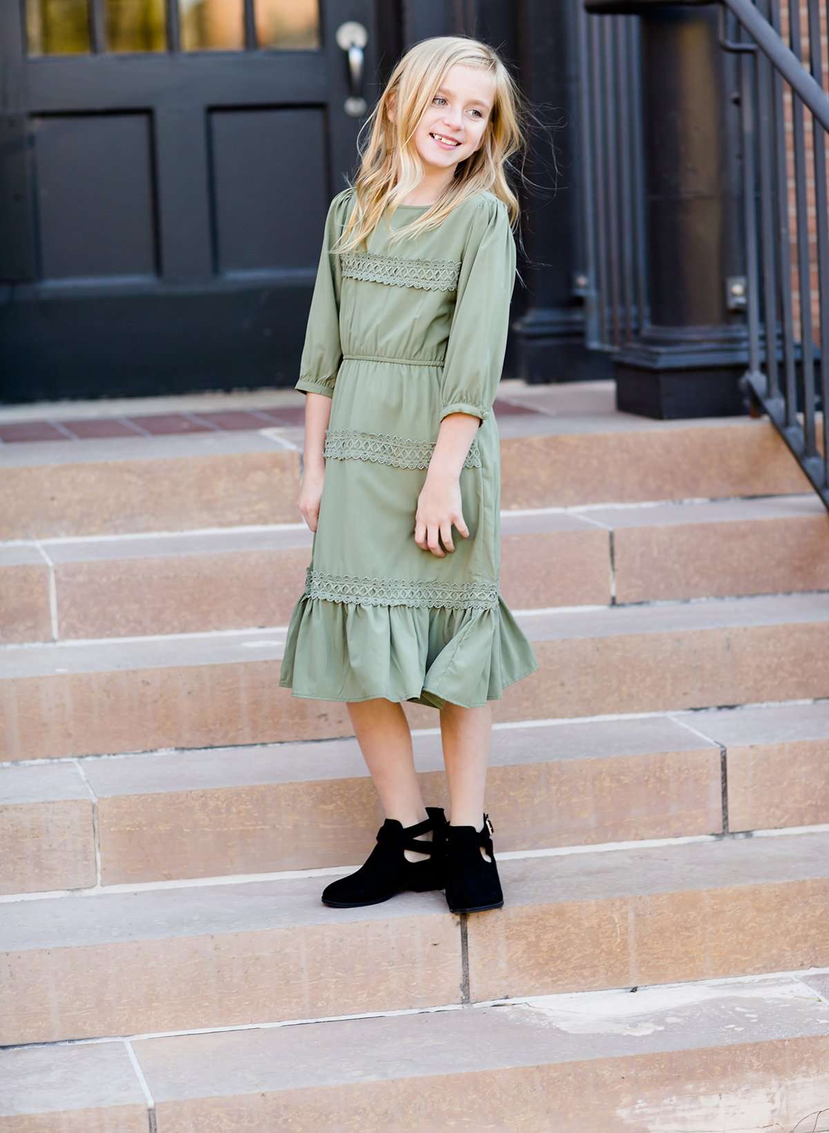 Young girl wearing a sage colored ruffle dress with lace trim on the tiers of the ruffles.